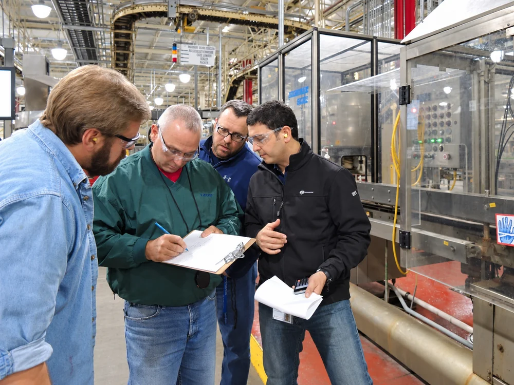 Four team members wearing safety glasses review notes on a clipboard beside guarded production equipment.
